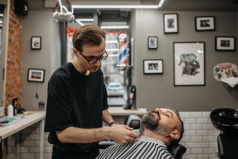 A barber meticulously trims a bearded man's hair in a contemporary barbershop setting.